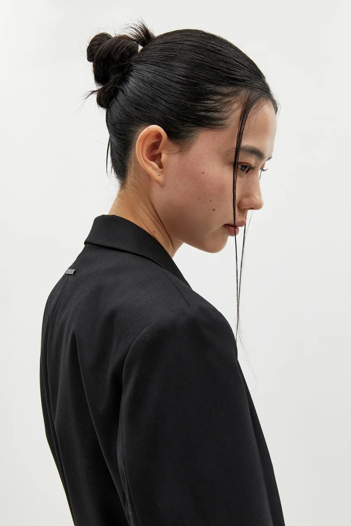 Side profile of a woman with sleek bun hairstyle wearing a black wool blazer against white background