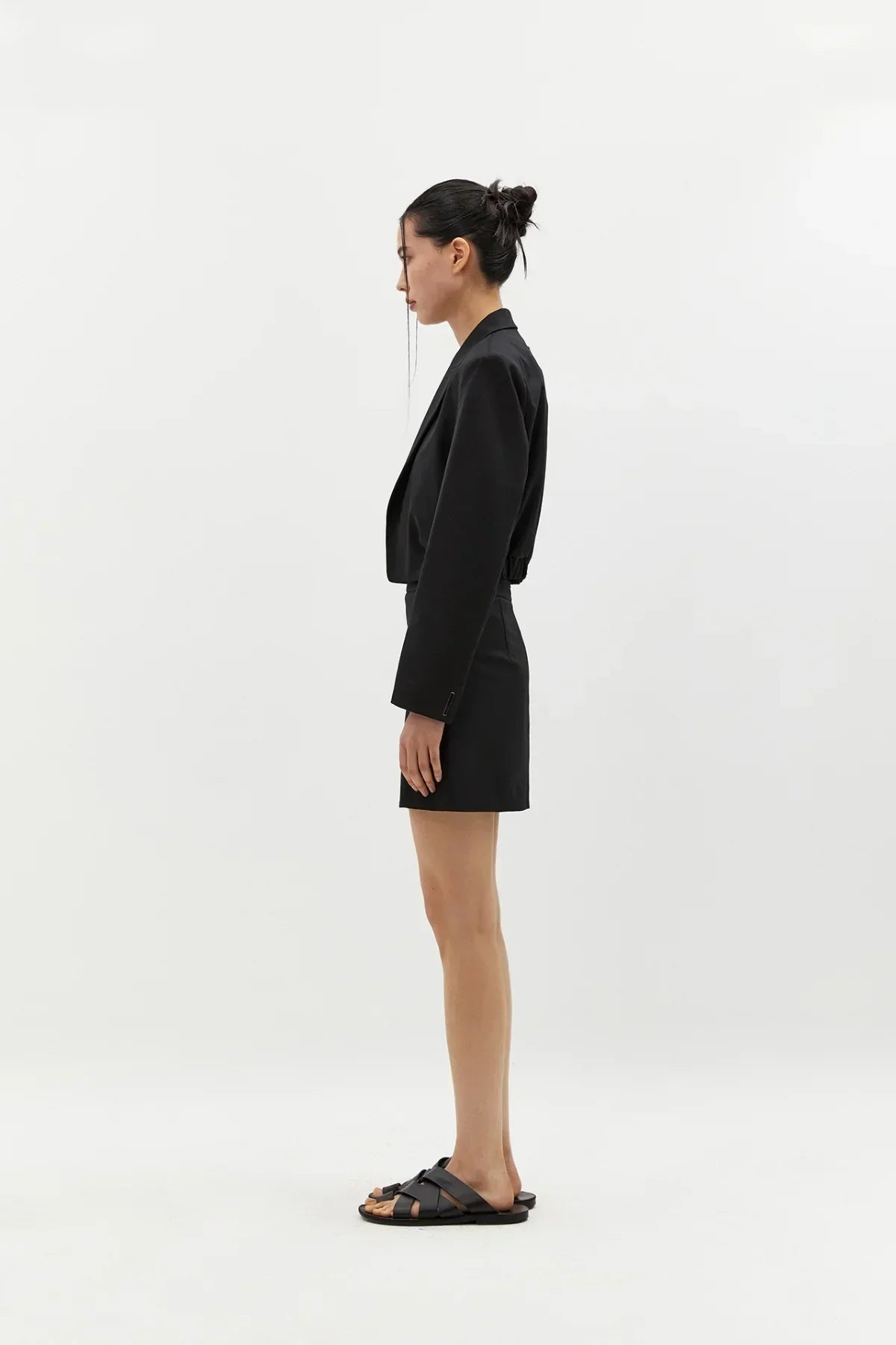 Side view of a woman wearing a black cropped wool blazer, matching skirt, and black sandals on white background