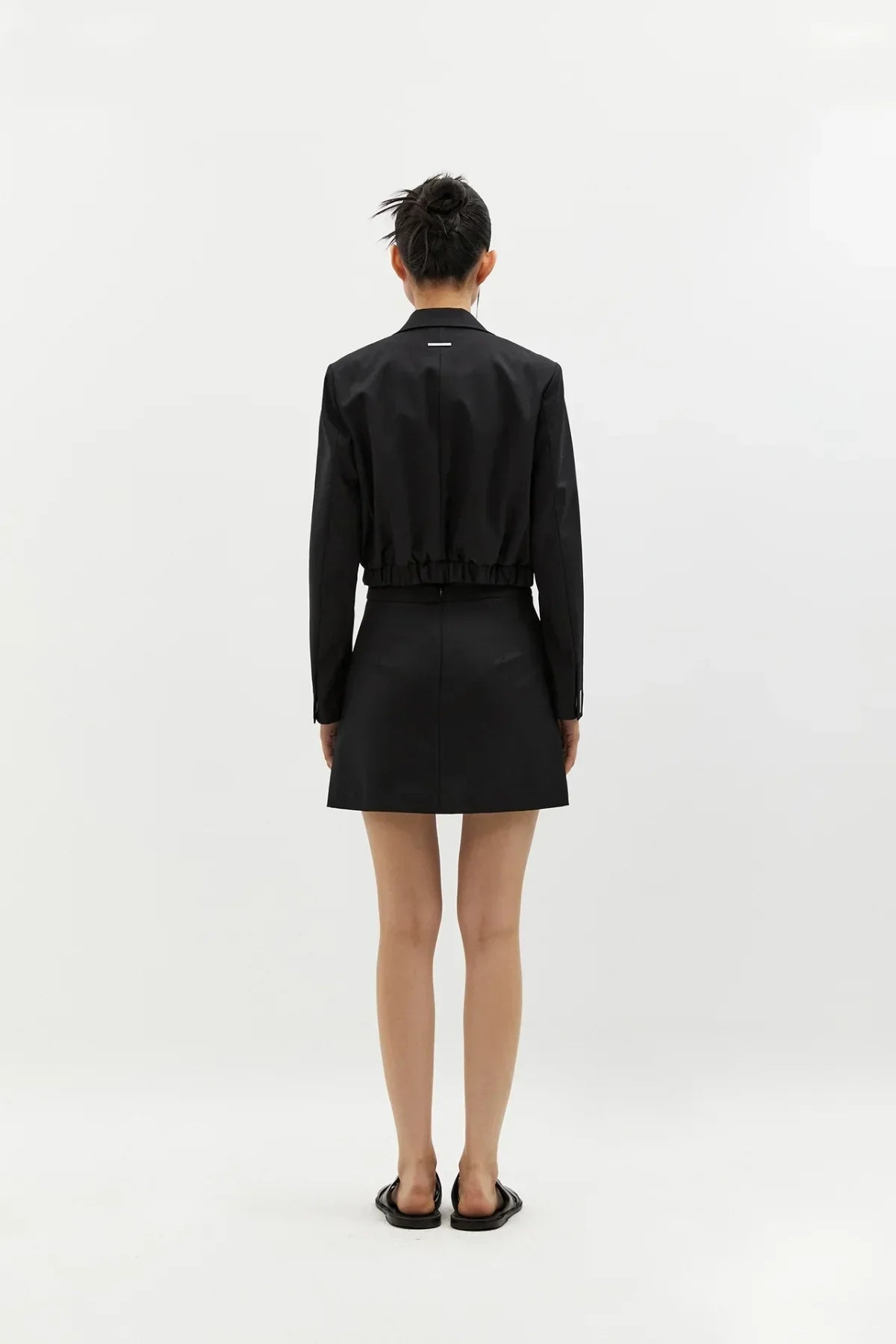 Back view of model wearing black cropped wool blazer and black mini skirt with black slip-on shoes