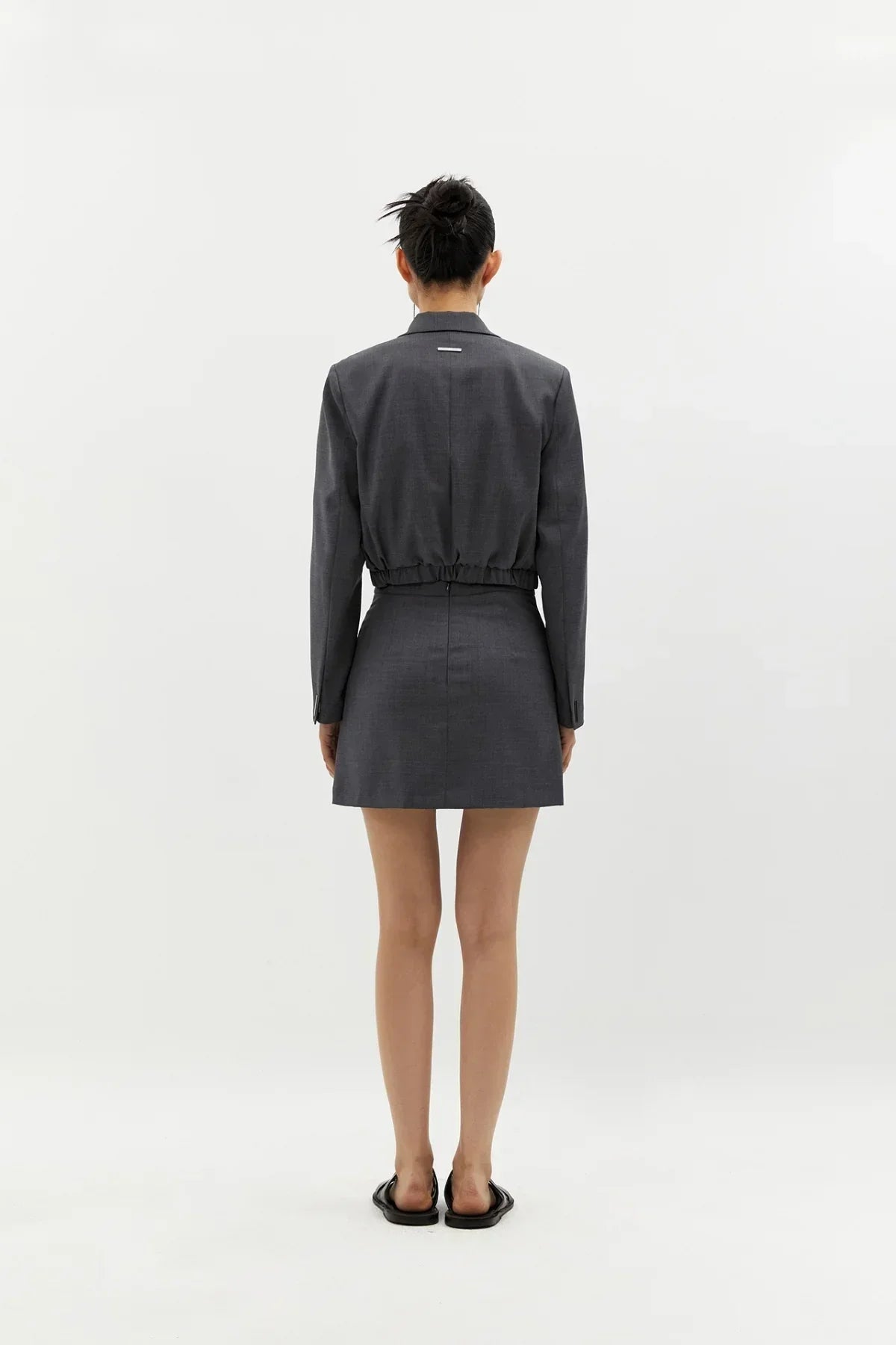 Rear view of model wearing grey wool cropped blazer and matching mini skirt with black flats