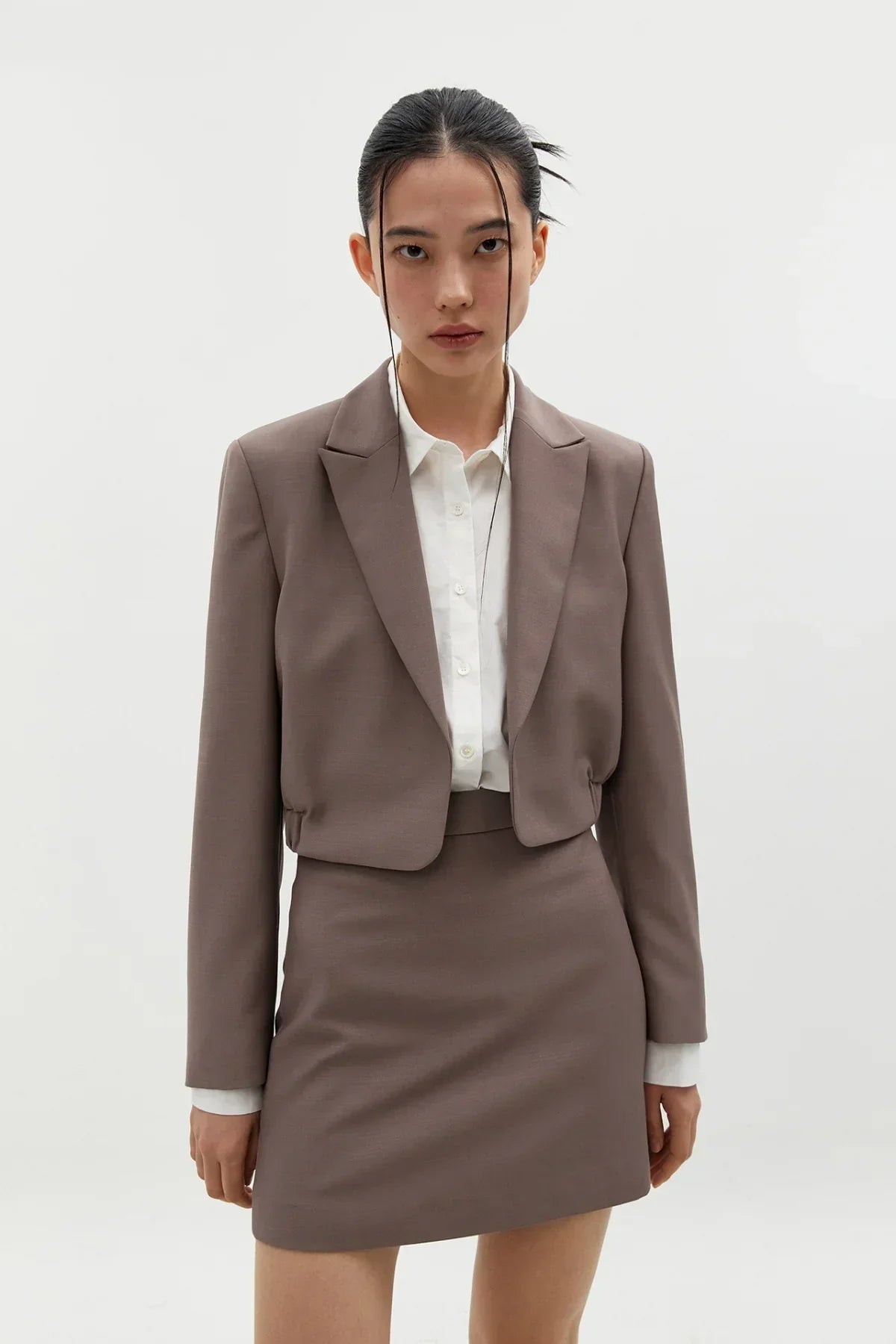 Model wearing taupe cropped blazer and matching skirt with white button-up shirt in studio