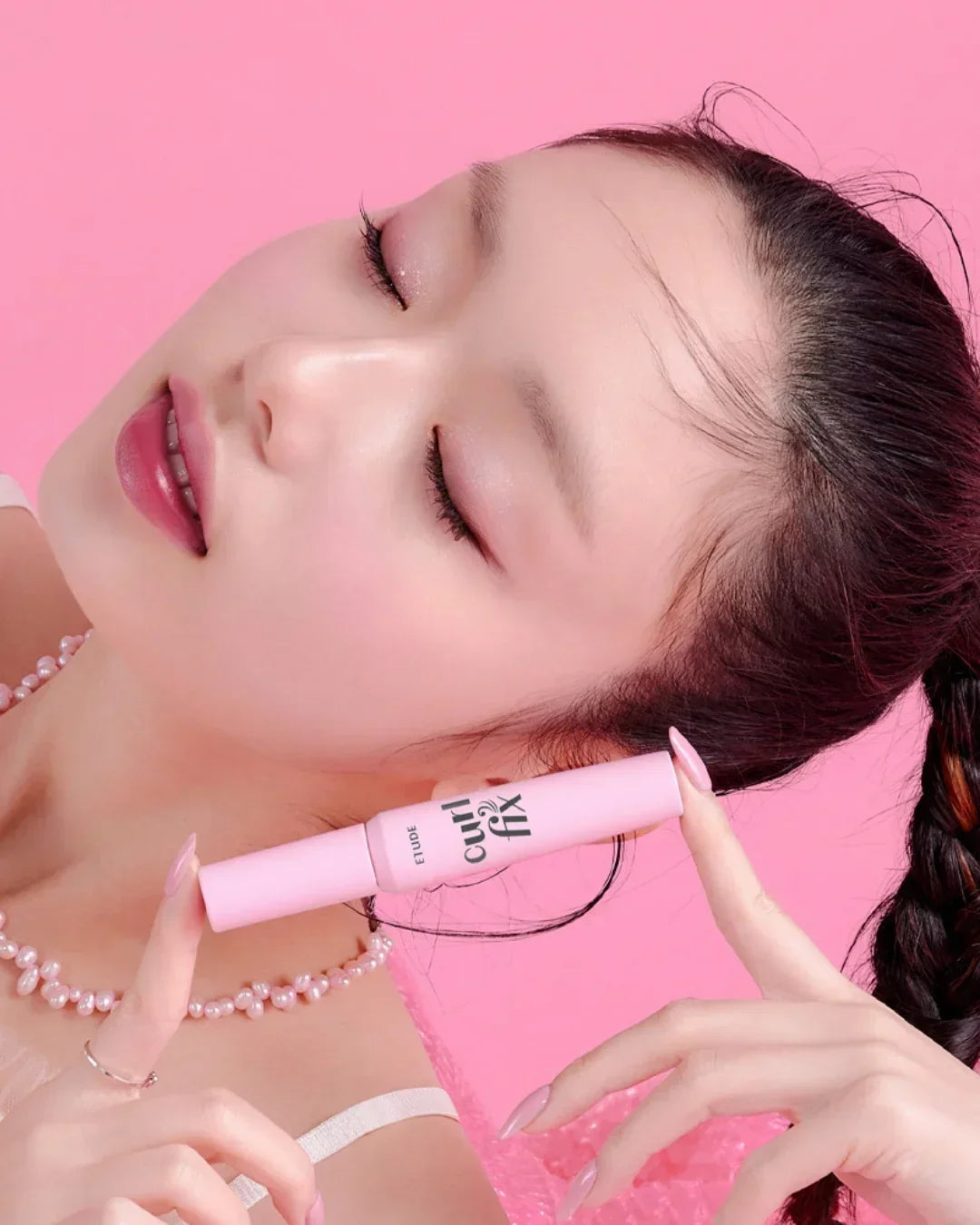 Close-up of woman with braided hair holding pink Etude Curl Fix mascara against a pink background