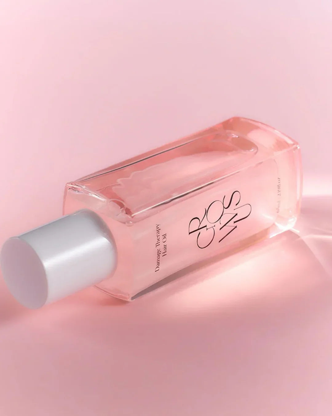 Clear rectangular bottle of damage therapy hair oil with white cap on pink background