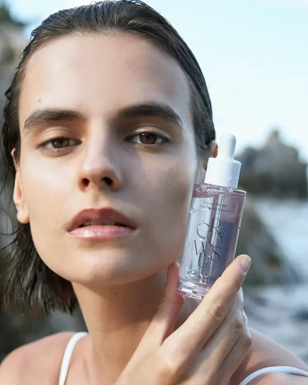 Close-up of a woman holding a transparent skincare ampoule bottle labeled CROWSUS against an outdoor blurred background