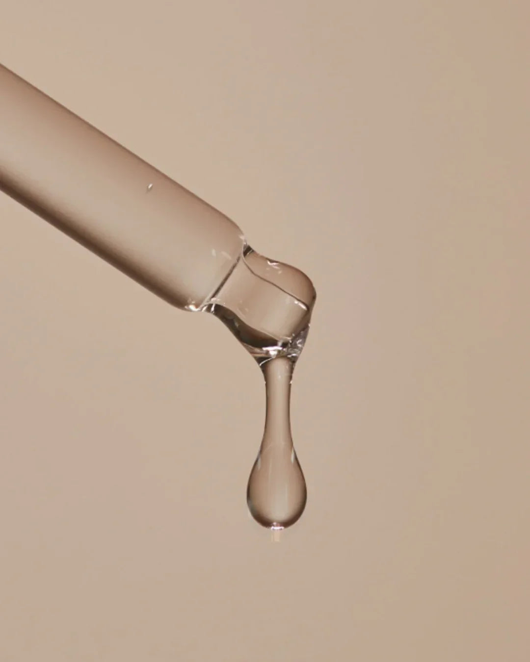 Close-up of clear dropper with a drop of liquid serum against a beige background