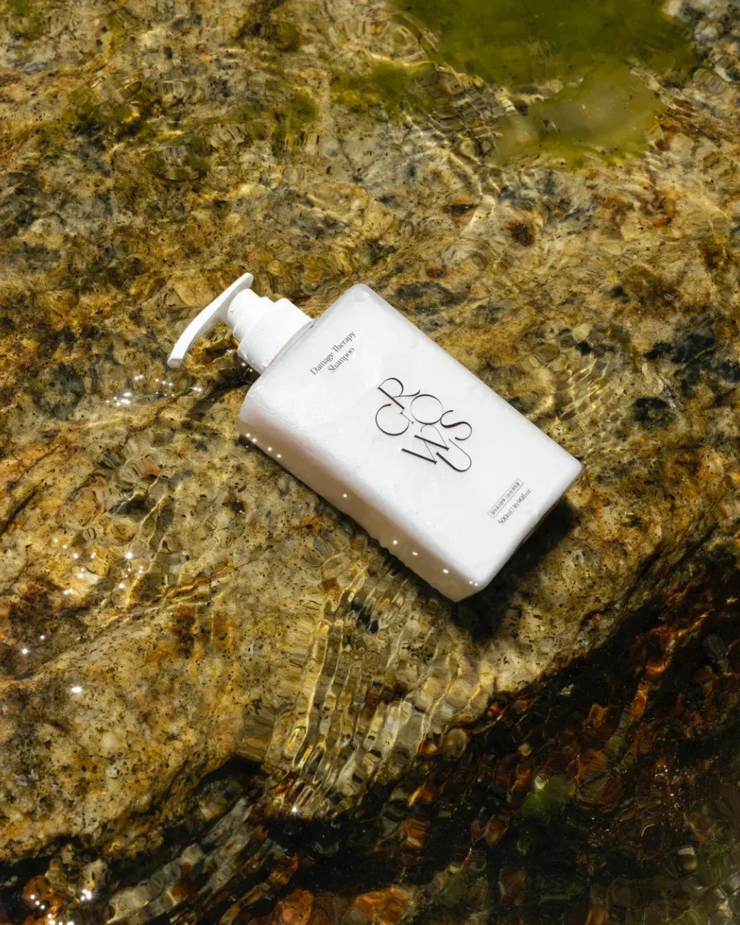 White bottle of damage therapy shampoo with pump resting on rocky stream bed under clear water