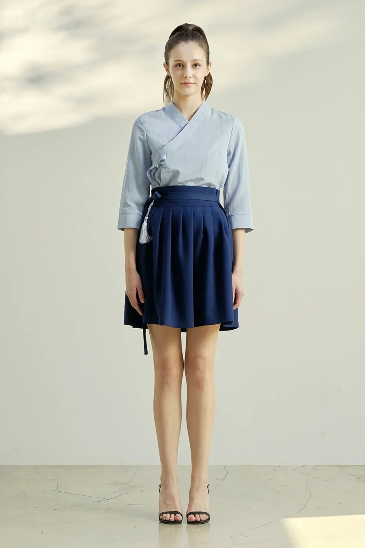 Woman wearing light blue wrap blouse and navy blue pleated mini skirt with tassel accessory
