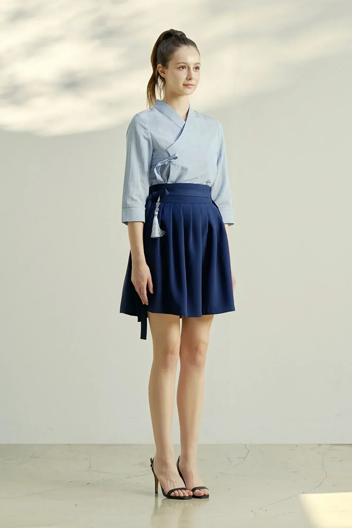 Woman wearing light blue wrap top and navy blue pleated mini skirt with tassel, black high heels