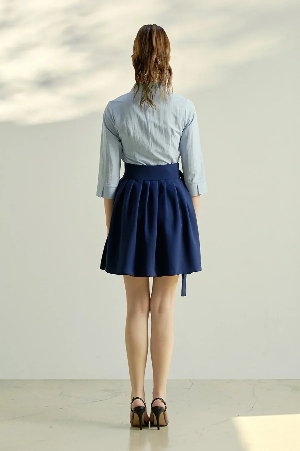 Woman model wearing a navy pleated mini skirt and a light blue three-quarter sleeve shirt, black high heels