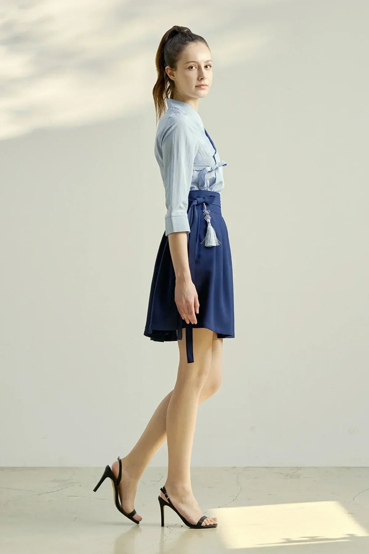 Young woman posing in studio wearing a light blue blouse and navy mini skirt with tassel detail