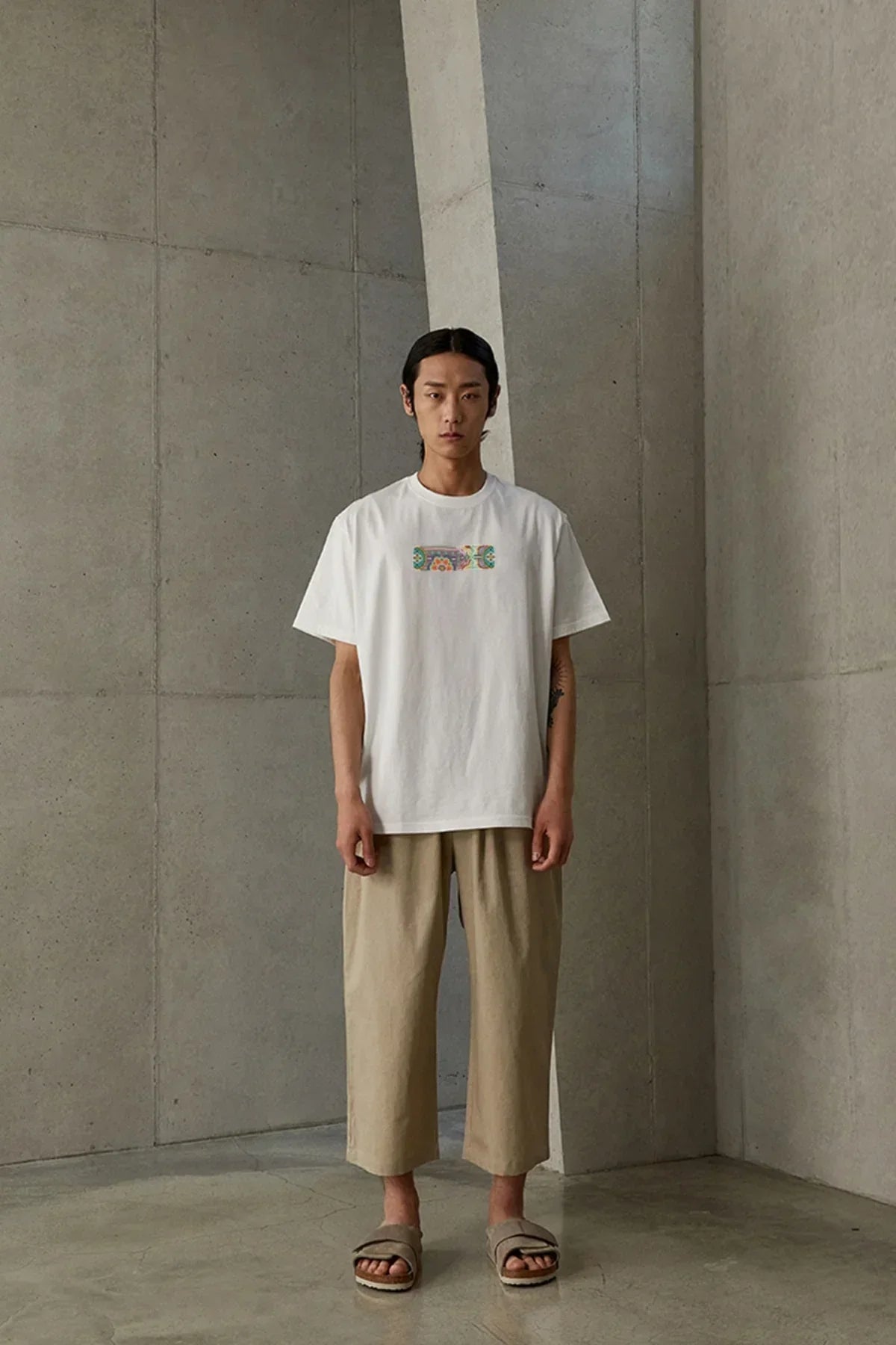Model wearing white Dancheong graphic t-shirt with beige cropped pants and sandals in minimalist concrete room