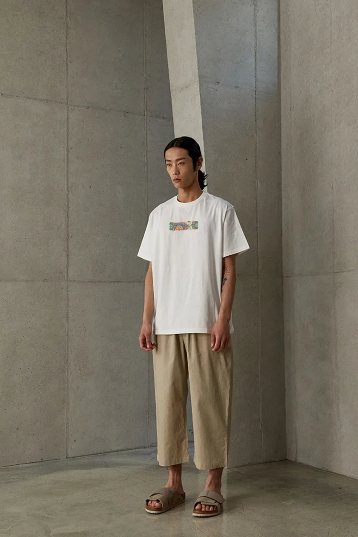 Man wearing white t-shirt with colorful print and beige cropped pants in minimalist concrete room