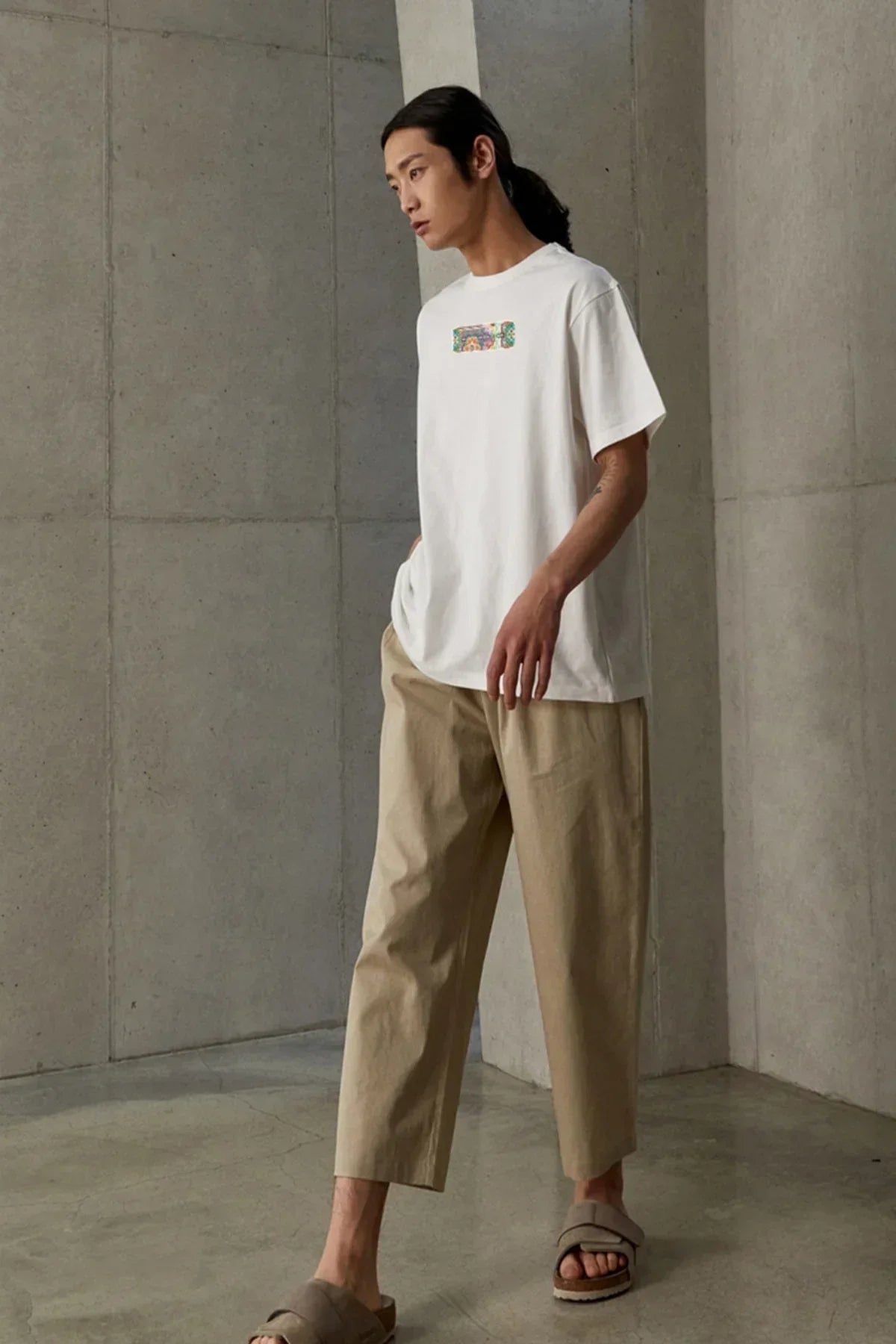 Male model in beige pants and white t-shirt with colorful rectangular graphic, standing in minimal concrete room