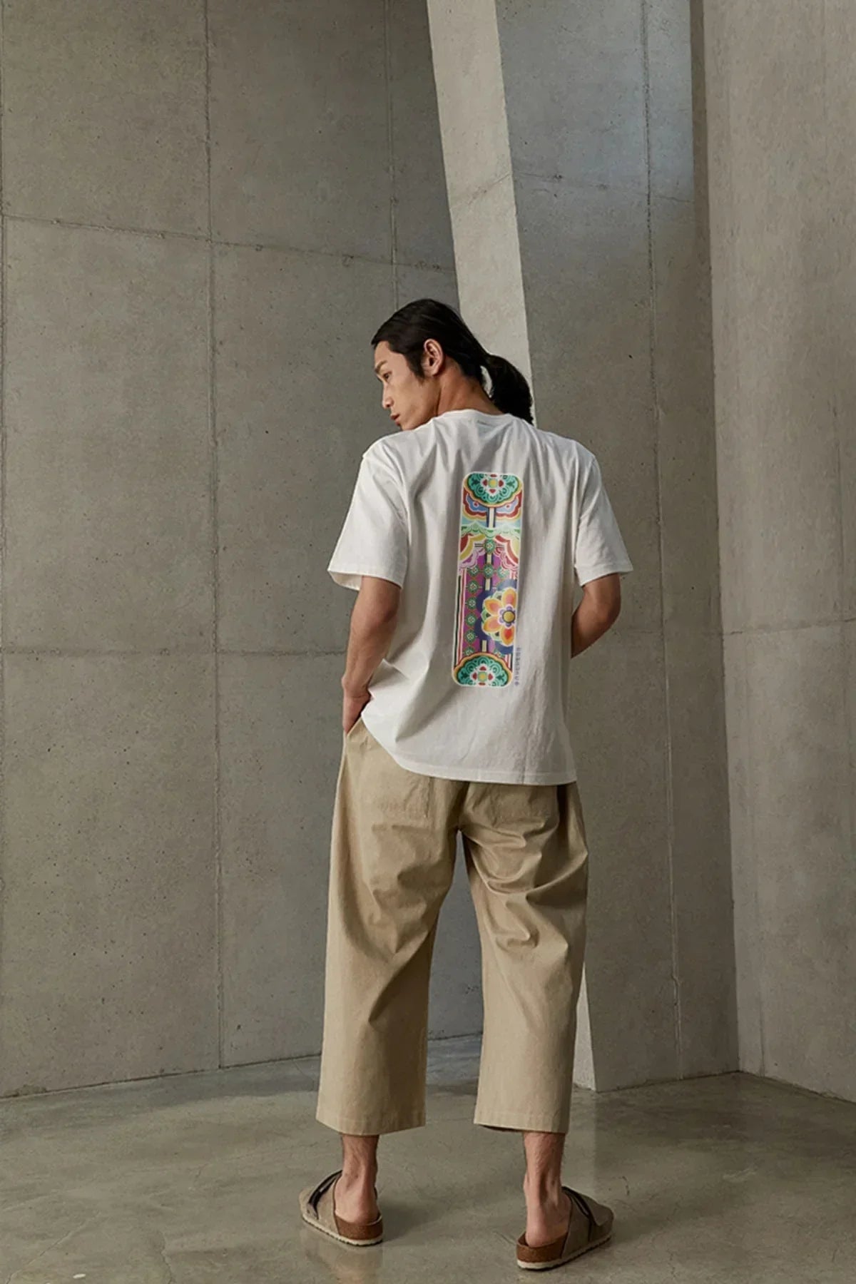 Man wearing white Dancheong T-shirt with colorful traditional Korean pattern on back, beige pants, minimal concrete interior