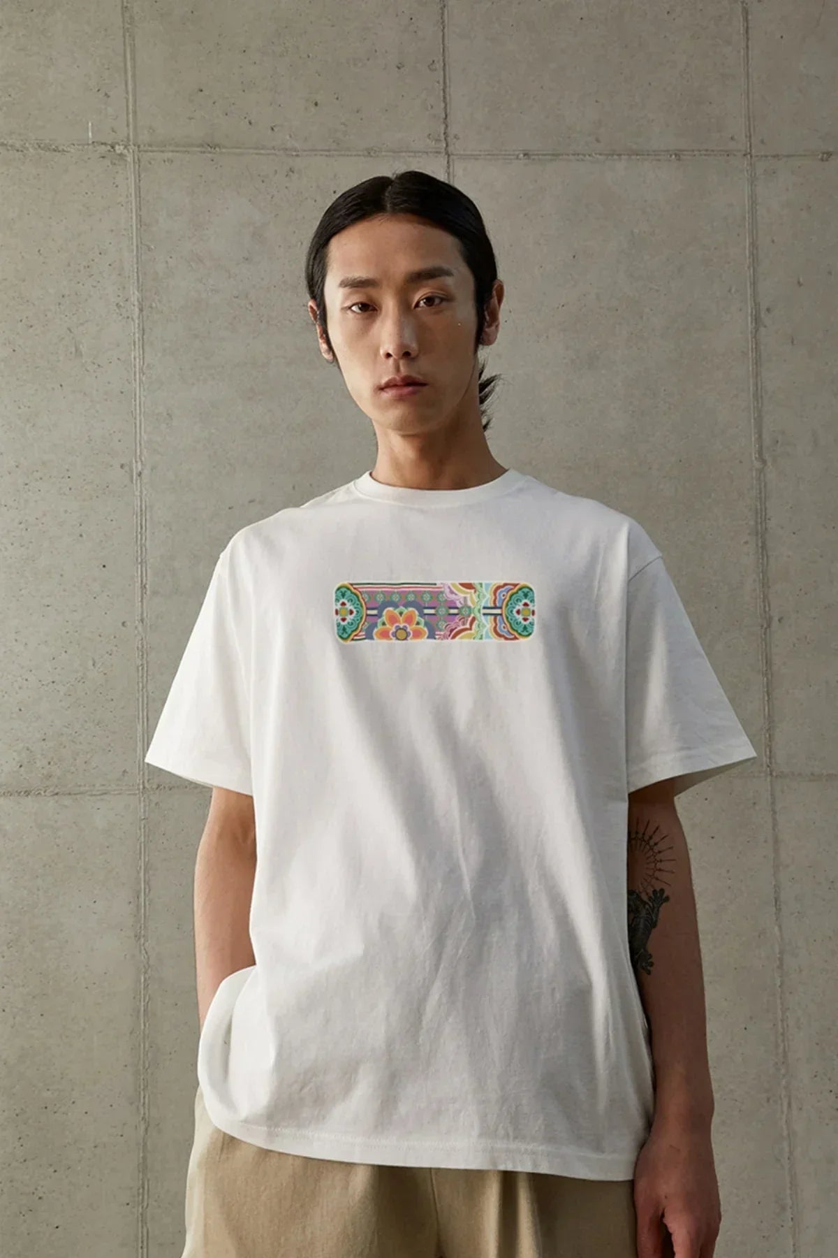 Man wearing white T-shirt with colorful Dancheong pattern print, standing against concrete wall