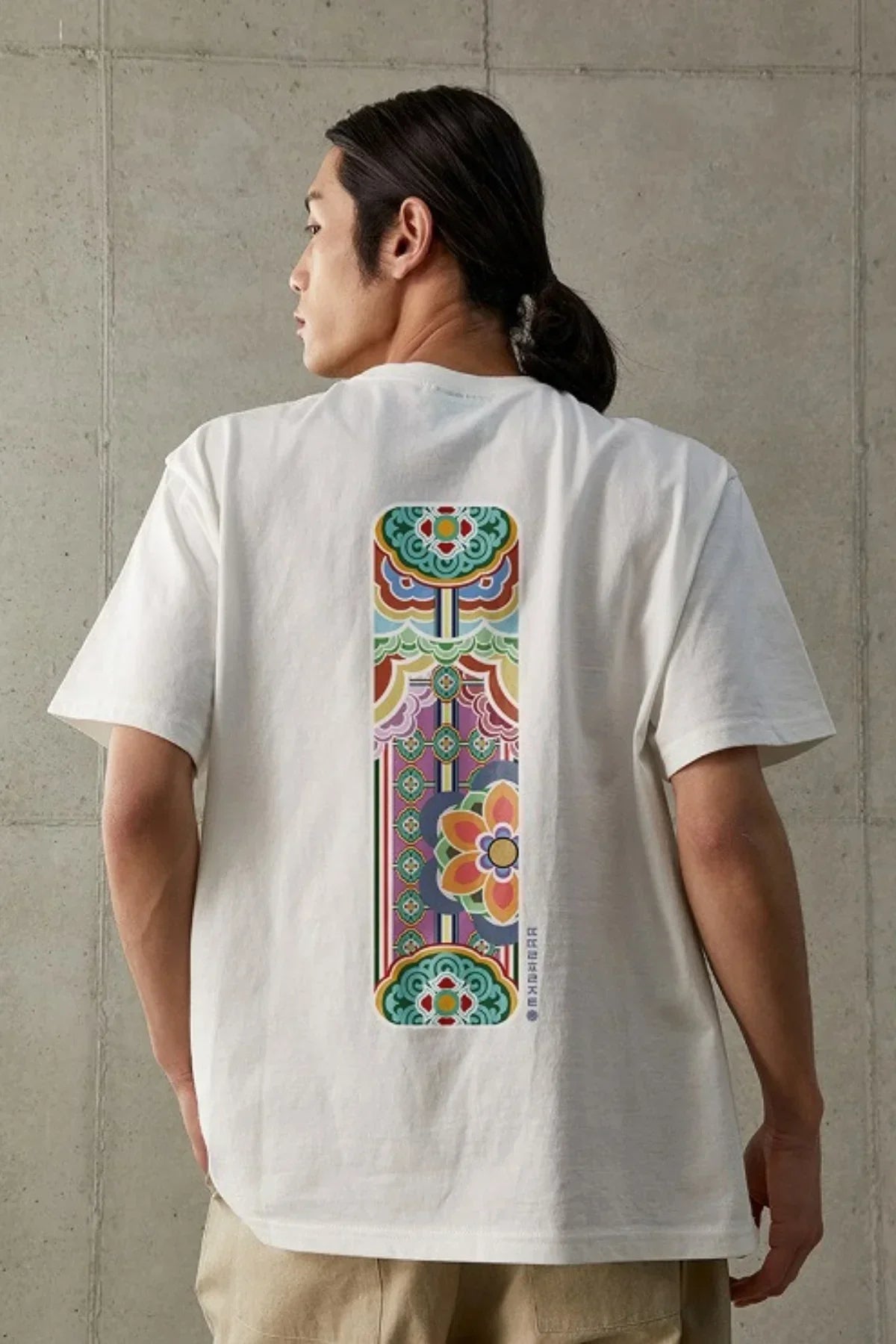 Man wearing white t-shirt with colorful traditional Dancheong pattern on back, standing against concrete wall