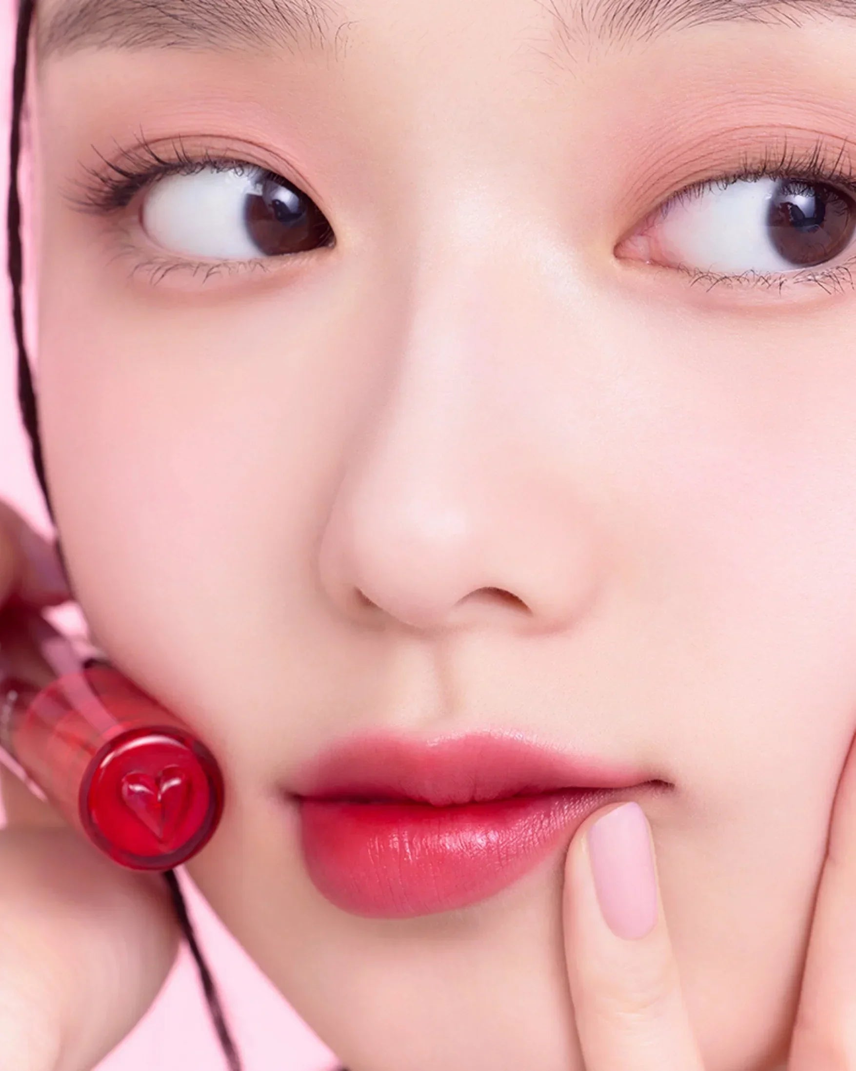 Close-up of woman's face with pink lips and holding red water gel lip tint against pink background