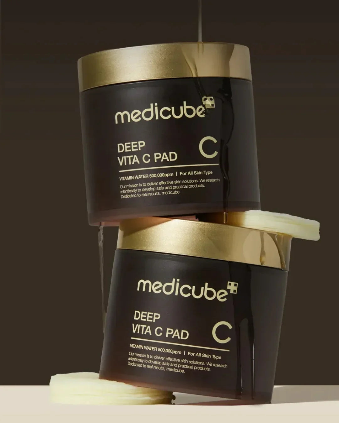 Two stacked Medicube Deep Vita C Pad containers with vitamin water dripping, cotton pads on beige surface