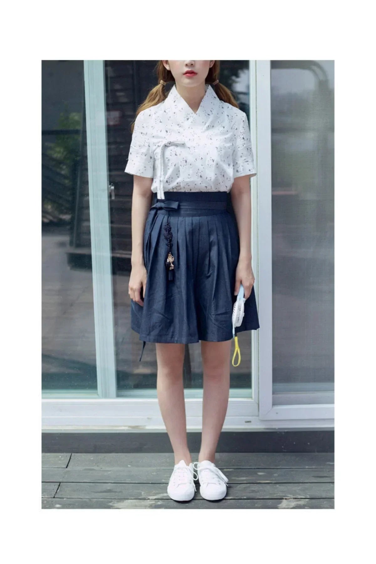 Woman wearing a white floral blouse and navy blue pleated skirt with white sneakers standing outdoors