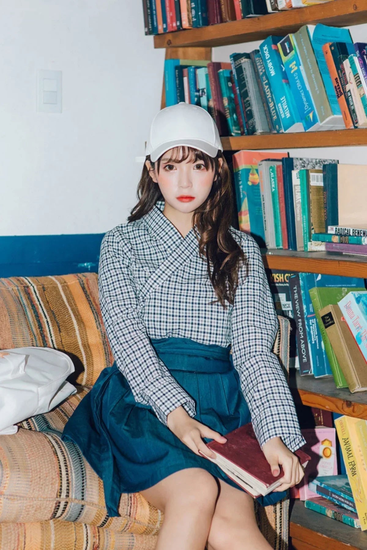 Young woman in white cap and checkered top sitting on couch holding book beside bookshelf