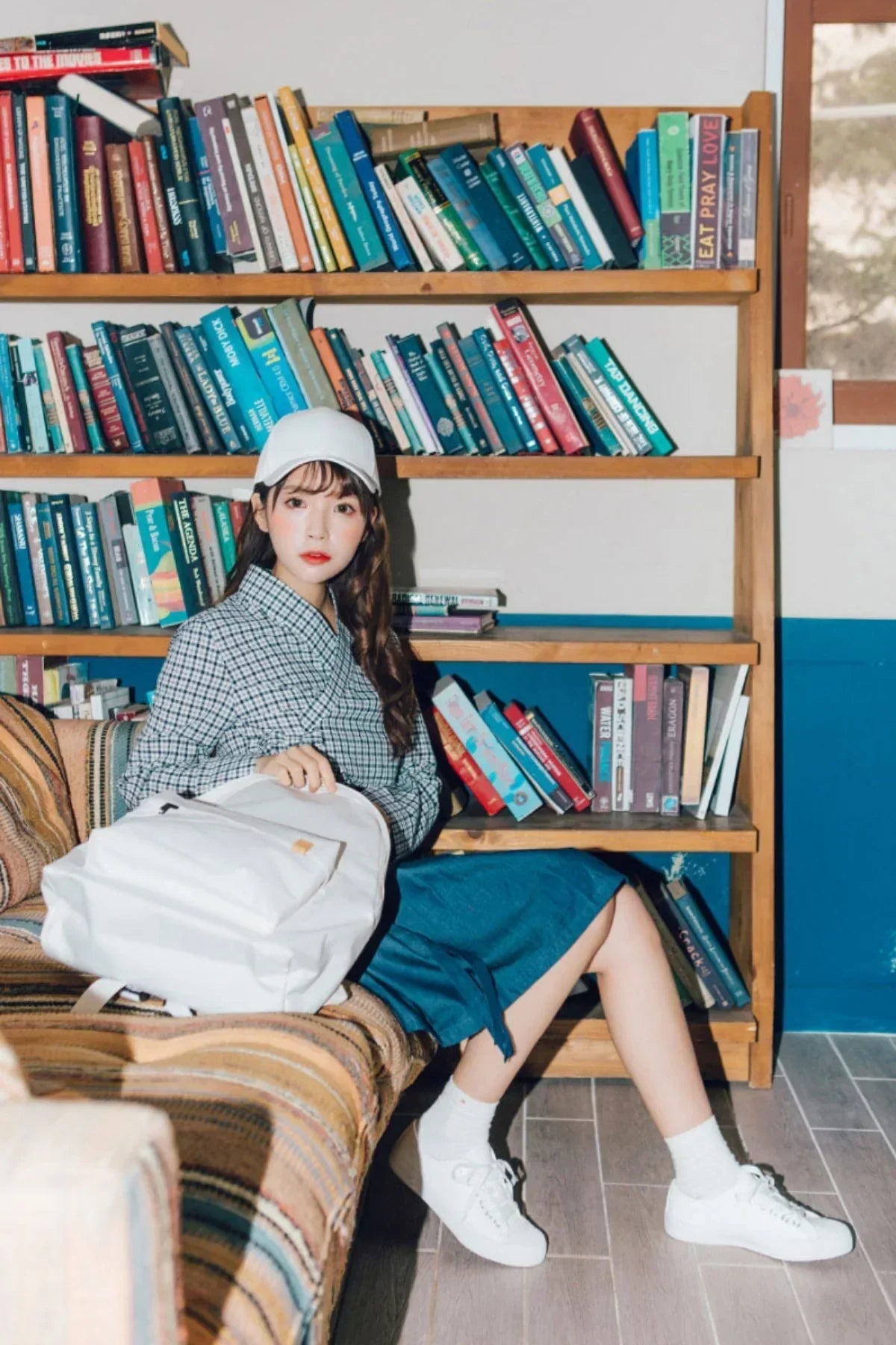 Young woman in checkered shirt, blue skirt, white cap, and sneakers sitting on a striped sofa with white backpack by bookshelf