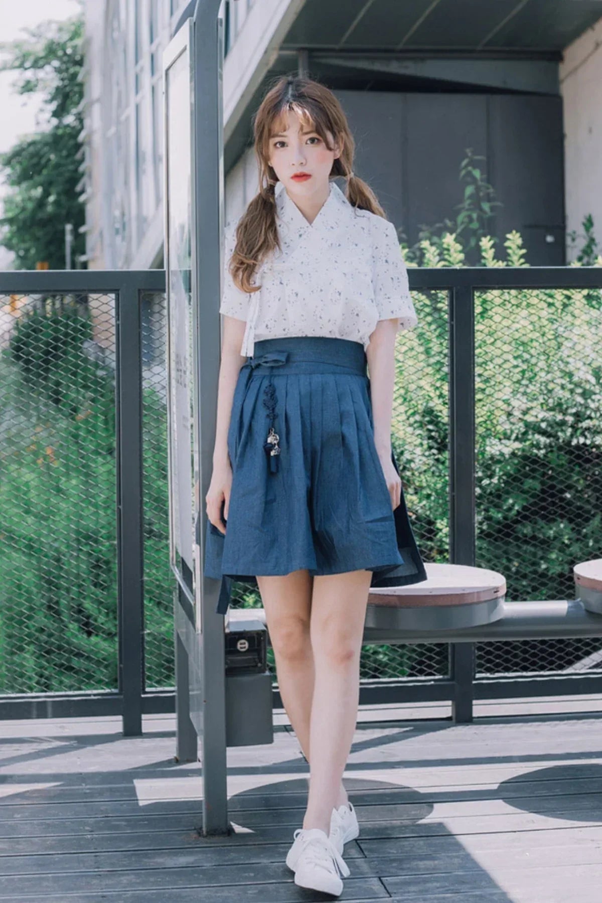 Young woman in a white floral blouse and blue pleated skirt standing by a modern metal fence outdoors