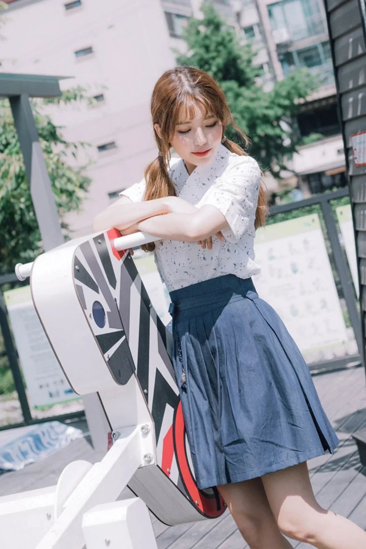 Young woman wearing a pleated navy blue skirt and white patterned blouse leaning on a colorful outdoor art installation