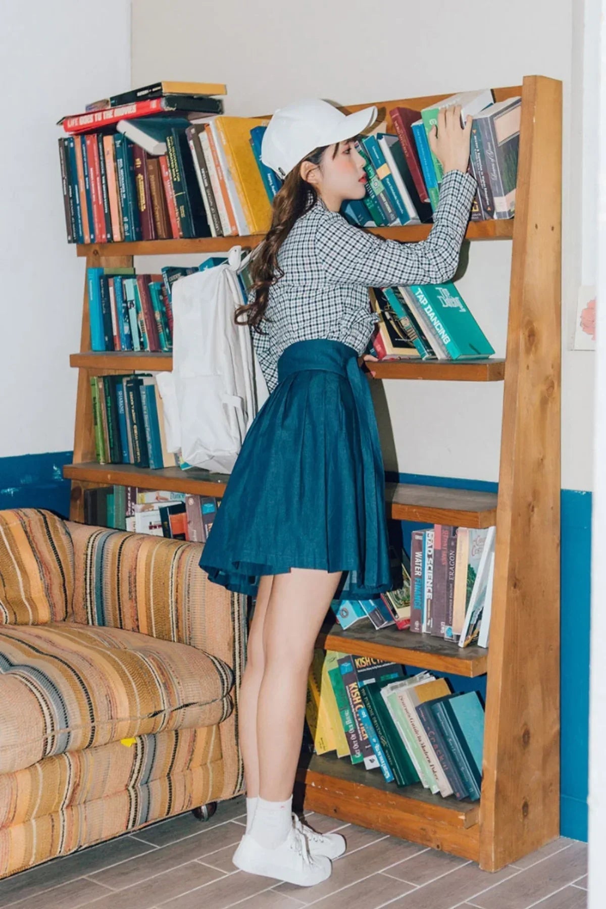 Young woman in white cap and blue skirt browsing books on a wooden bookshelf in cozy room