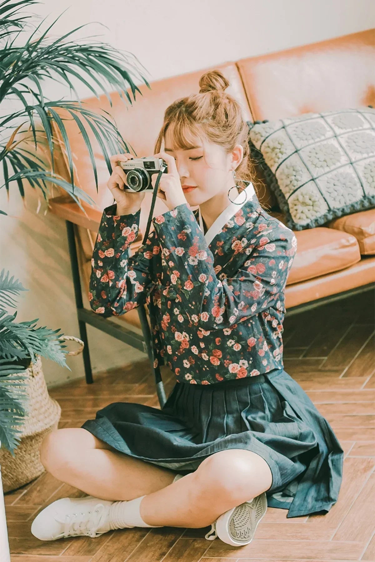 Young woman sitting on floor taking photo with vintage camera, wearing floral top and pleated skirt