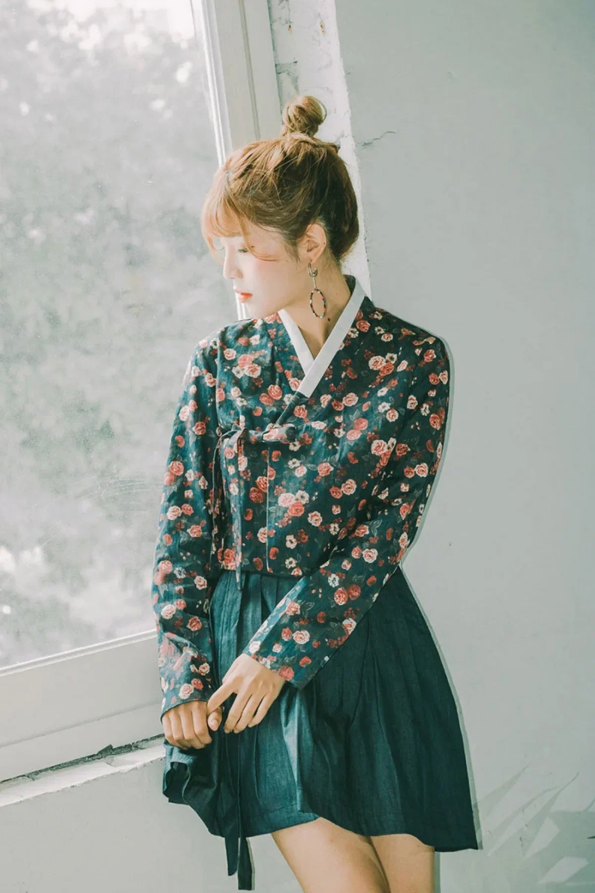 Woman wearing floral patterned hanbok top with dark pleated skirt standing by a window