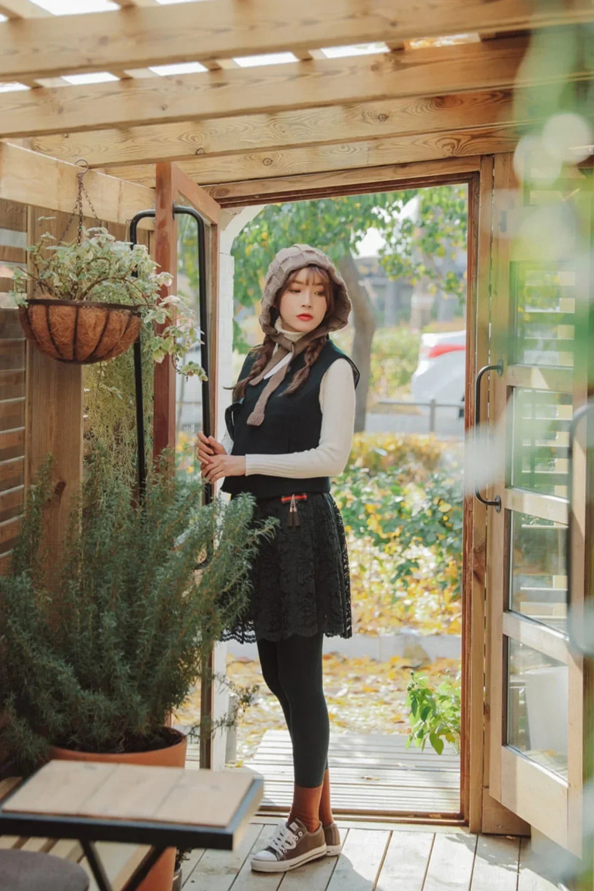 Woman wearing a black lace mini skirt, beige sweater, vest, and knitted bonnet standing in a wooden porch doorway