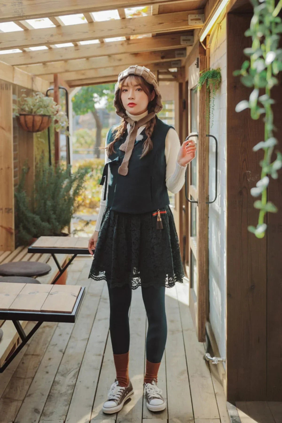 Young woman in black lace mini skirt, vest, and knitted hat standing on wooden porch with greenery