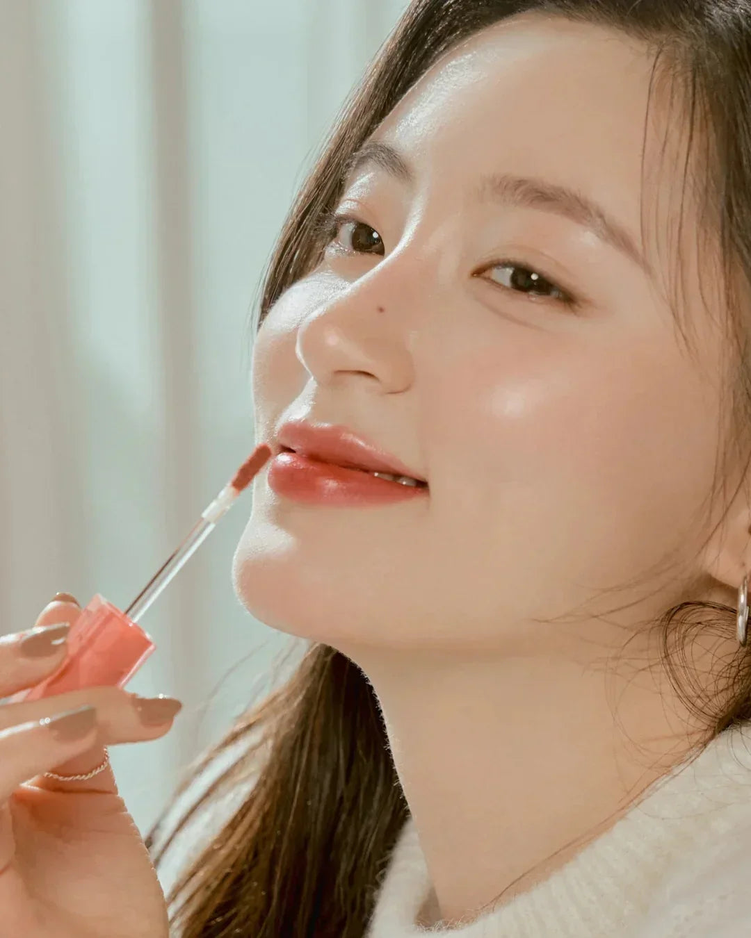 Close-up of woman applying dewy lip tint with applicator, glowing skin, natural makeup look