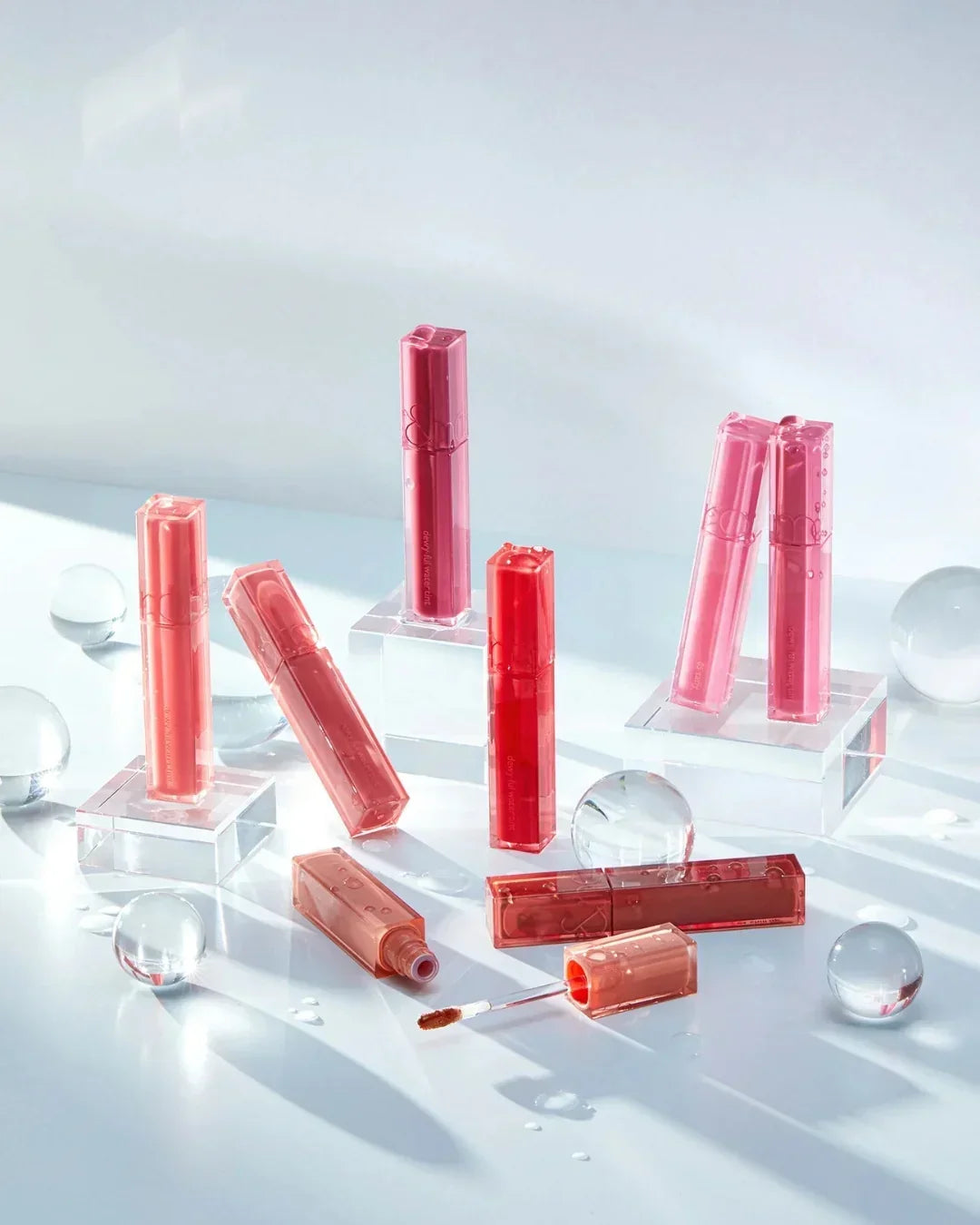 Dewyful Water Tint lip glosses in various pink and red shades arranged on clear acrylic blocks with water droplets and glass spheres