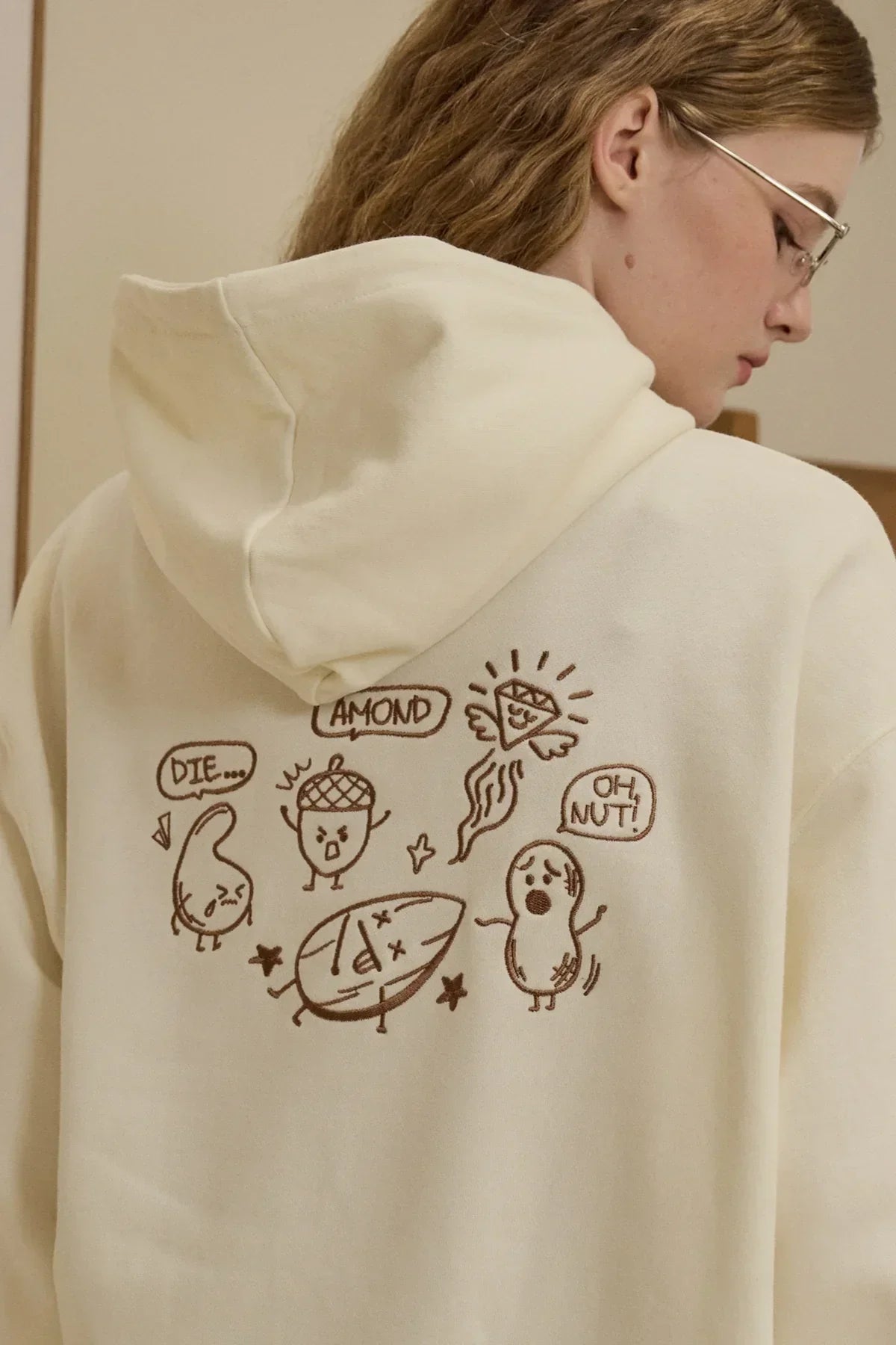 Close-up of cream hoodie with playful brown nut characters graphic on back, worn by woman with glasses
