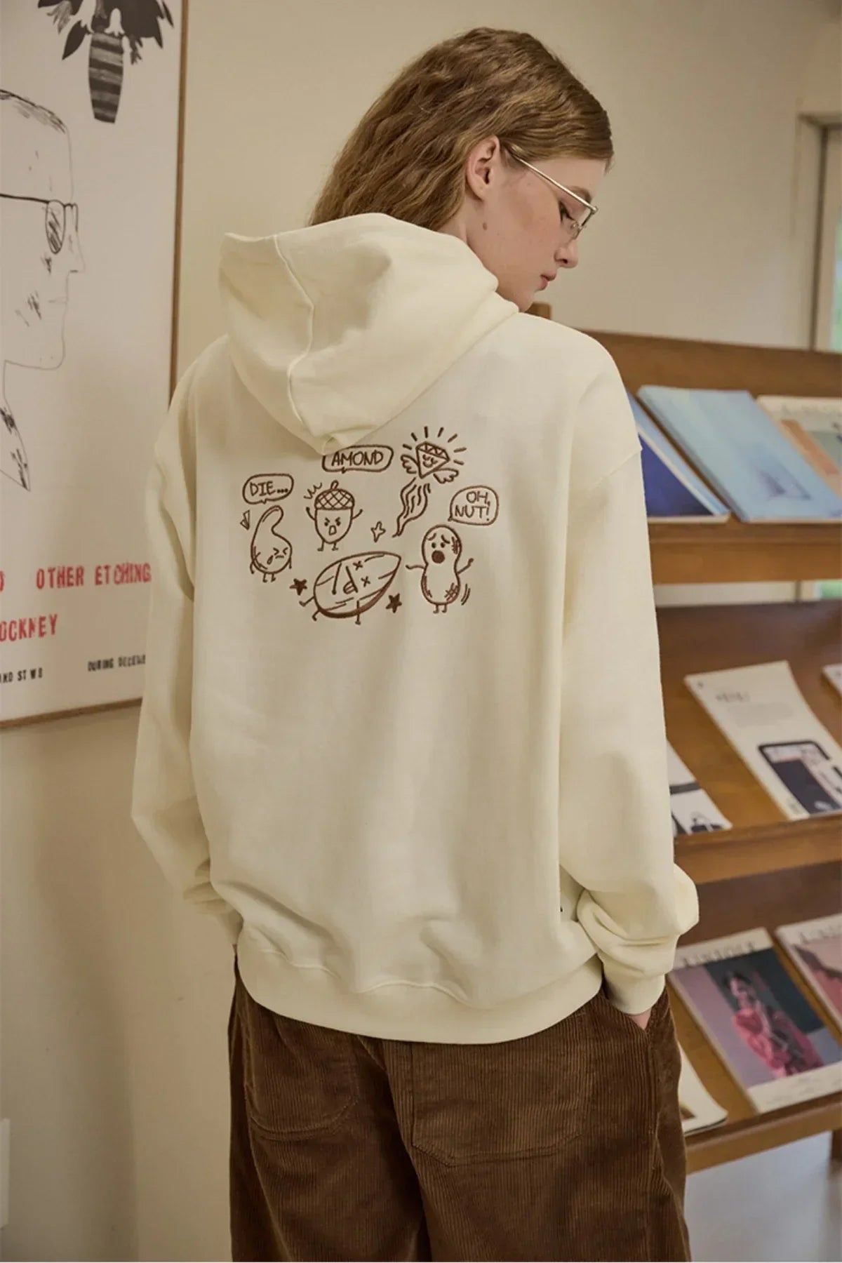 Person wearing cream hoodie with playful nut-themed cartoon embroidery on back in indoor setting