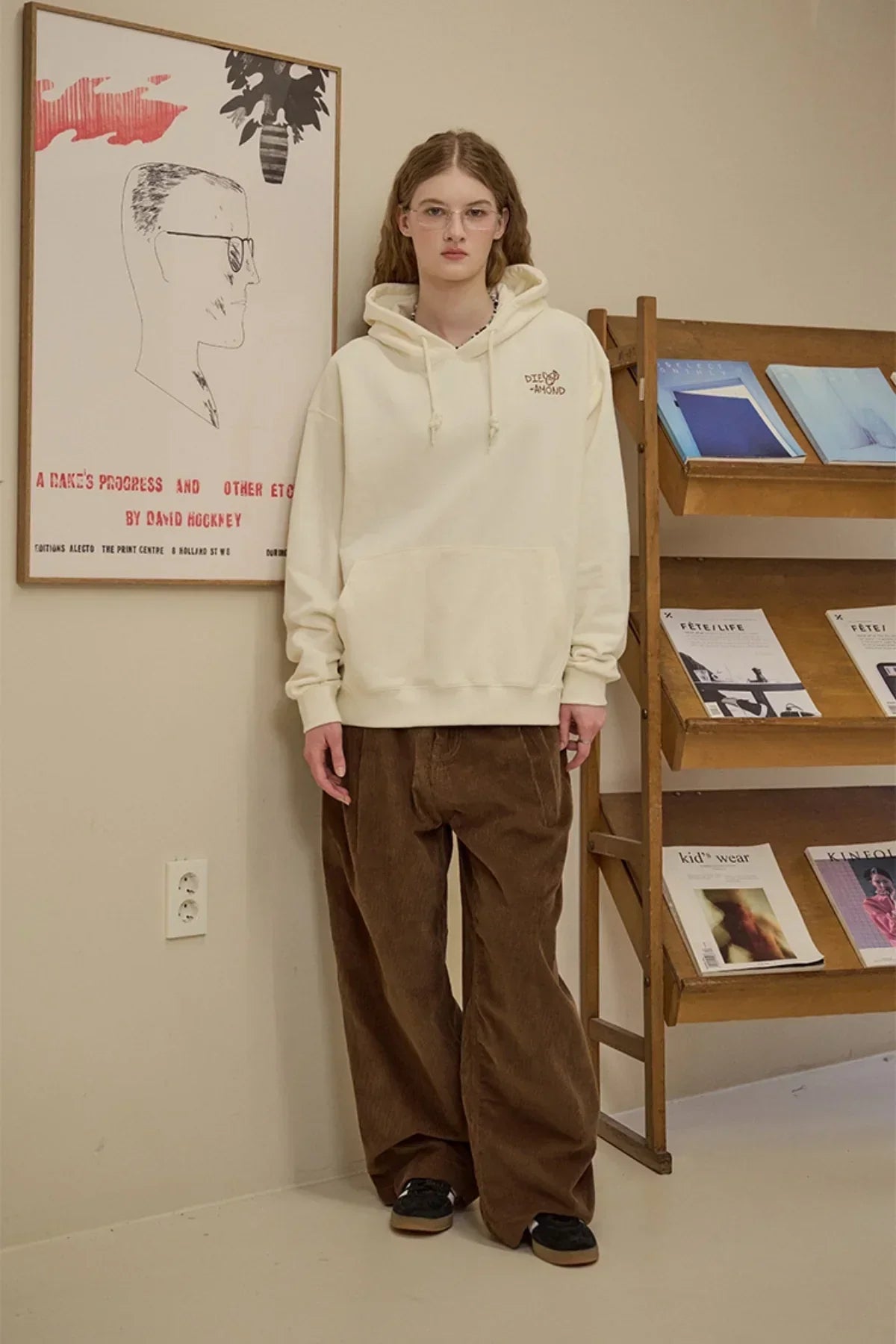 Model wearing cream diamond embroidered hoodie and brown wide-leg pants standing indoors near wooden magazine rack