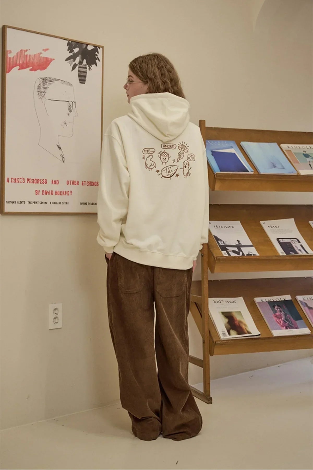 Woman wearing cream hoodie with cartoon print on back and loose brown corduroy pants indoors