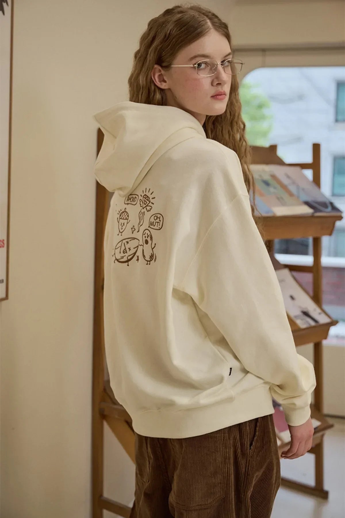 Young woman wearing cream hoodie with cartoon prints on back and brown corduroy pants in cozy indoor setting