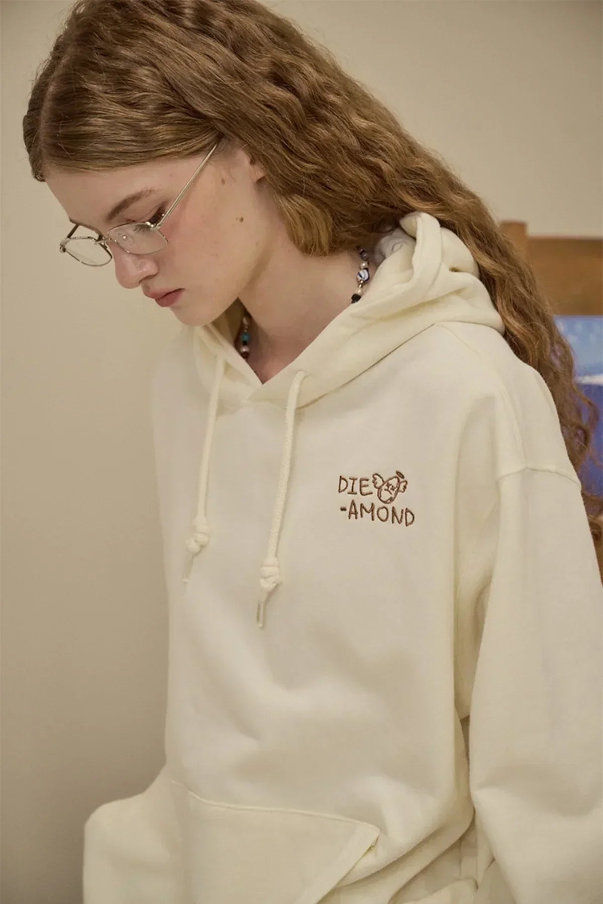 Young woman with glasses wearing cream hoodie with diamond embroidery in a neutral studio setting