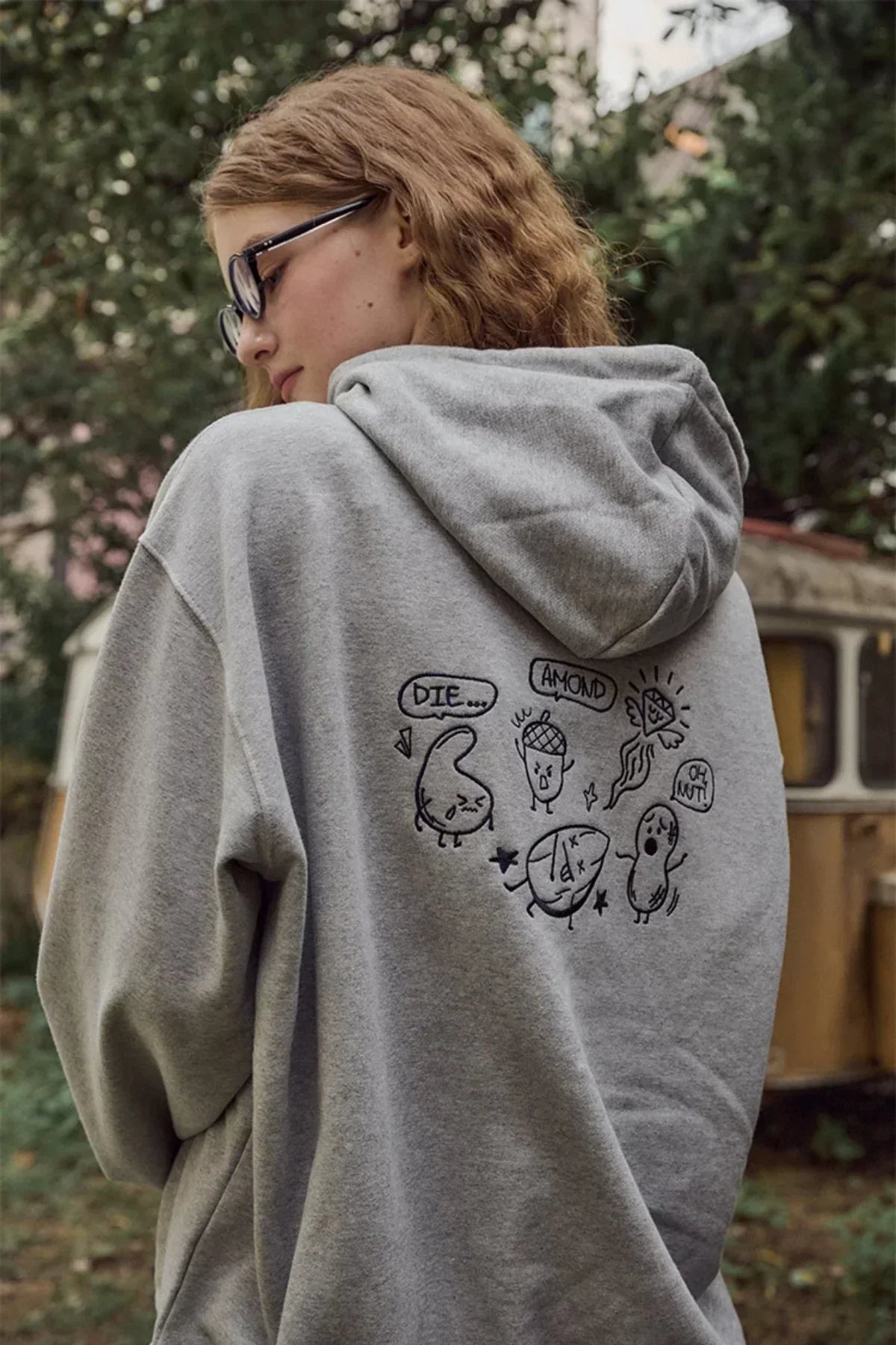 Woman in gray hoodie with cartoon nut characters and speech bubbles outdoors near vintage camper