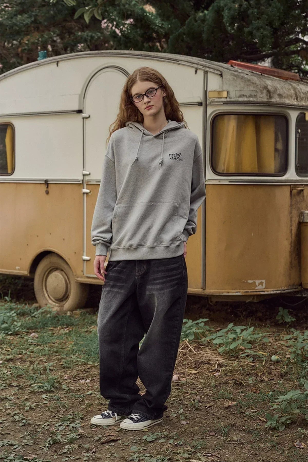 Young woman wearing gray Diamond embroidered hoodie and black baggy jeans standing outdoors by a vintage camper