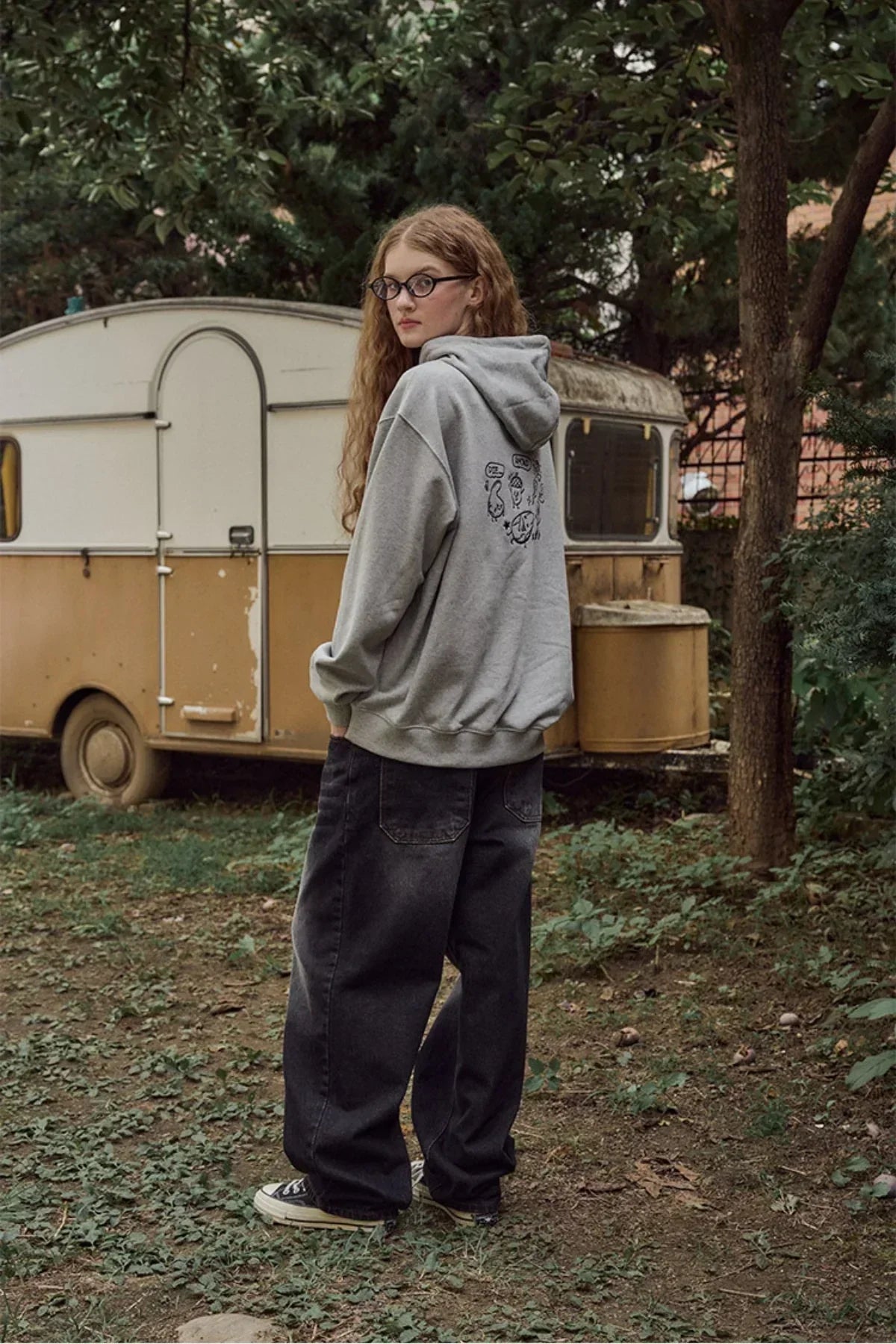 Young woman wearing gray hoodie with back print, loose black jeans, and sneakers outdoors near vintage yellow trailer