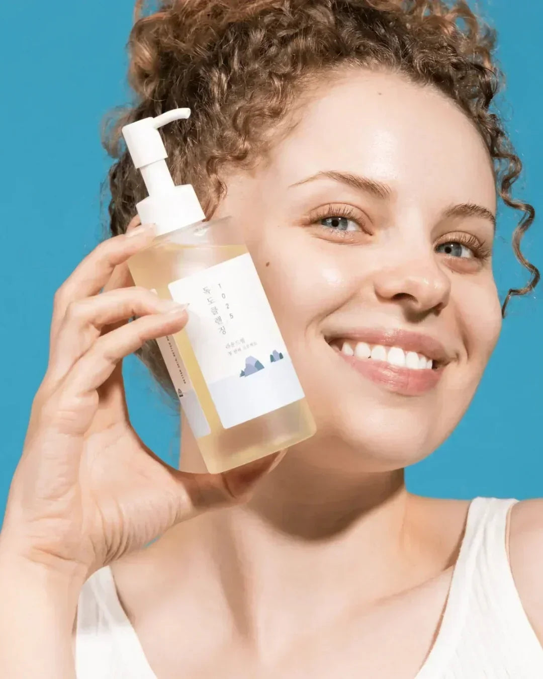 Smiling woman with curly hair holding Dokdo Cleansing Oil bottle against blue background