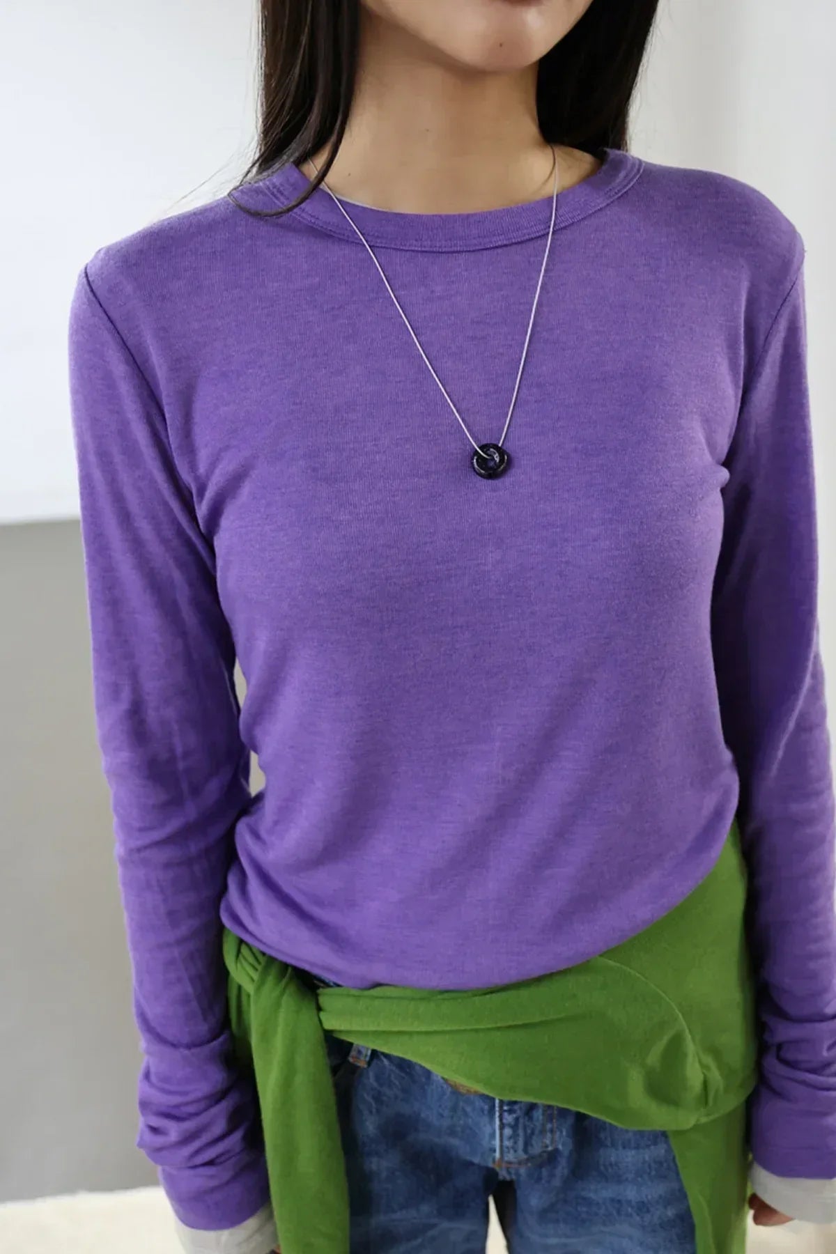 Close-up of woman wearing purple long-sleeve top, green sweater tied at waist, and black donut pendant necklace