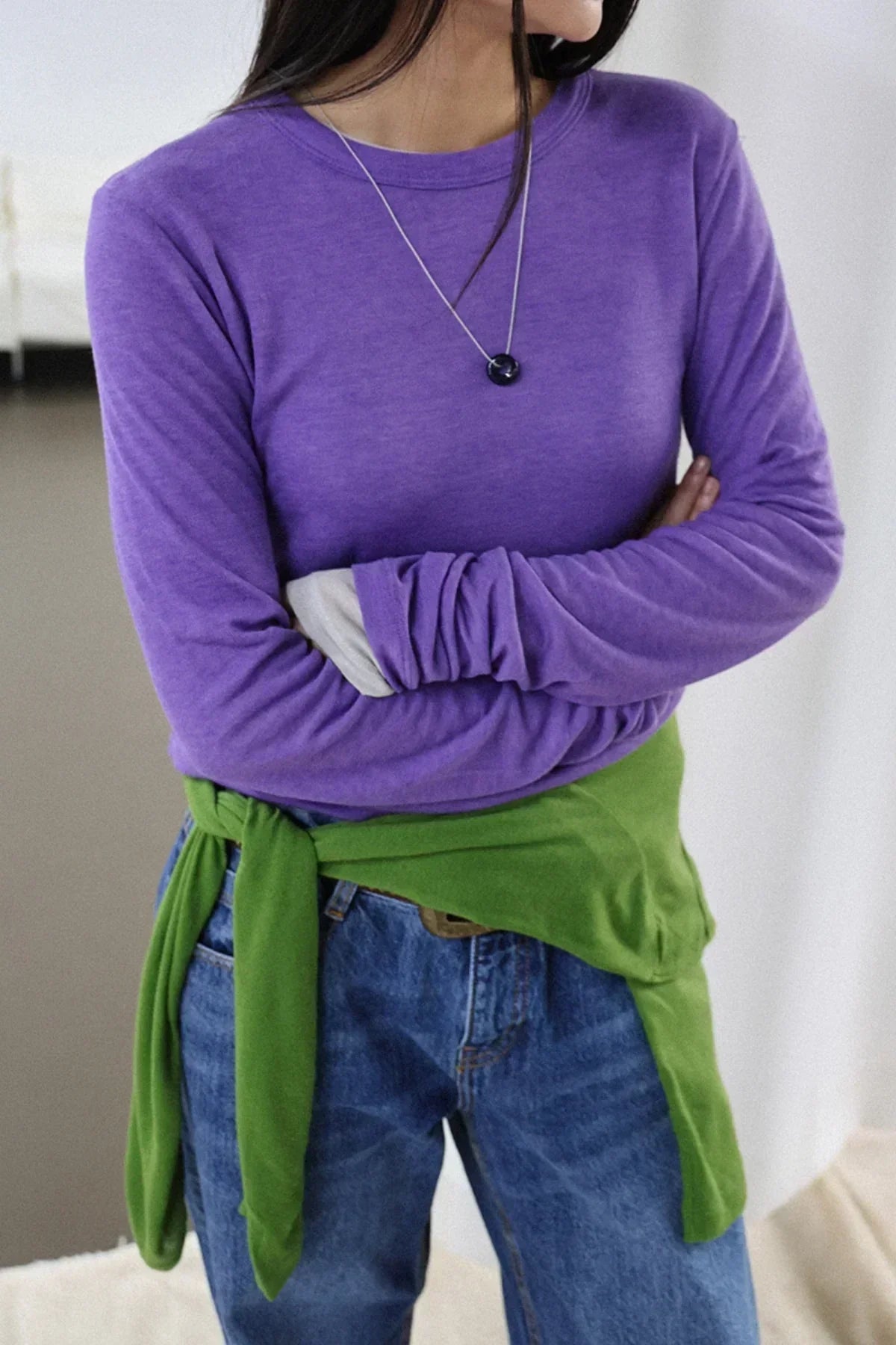 Close-up of person wearing purple long-sleeve shirt, blue jeans, green sweater tied around waist, and black donut necklace