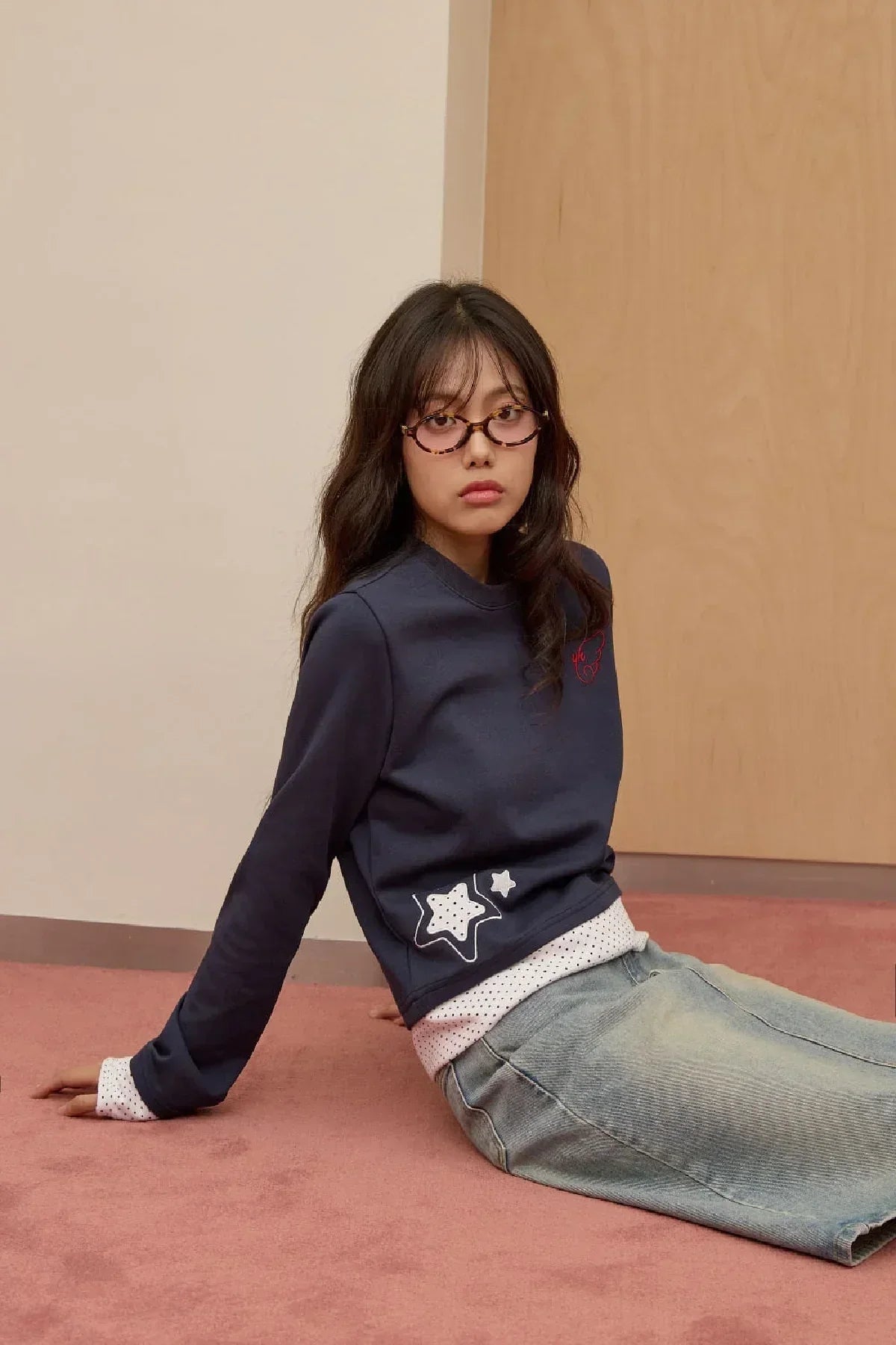 Young woman wearing navy star applique sweatshirt and light denim skirt sitting on pink carpet indoors