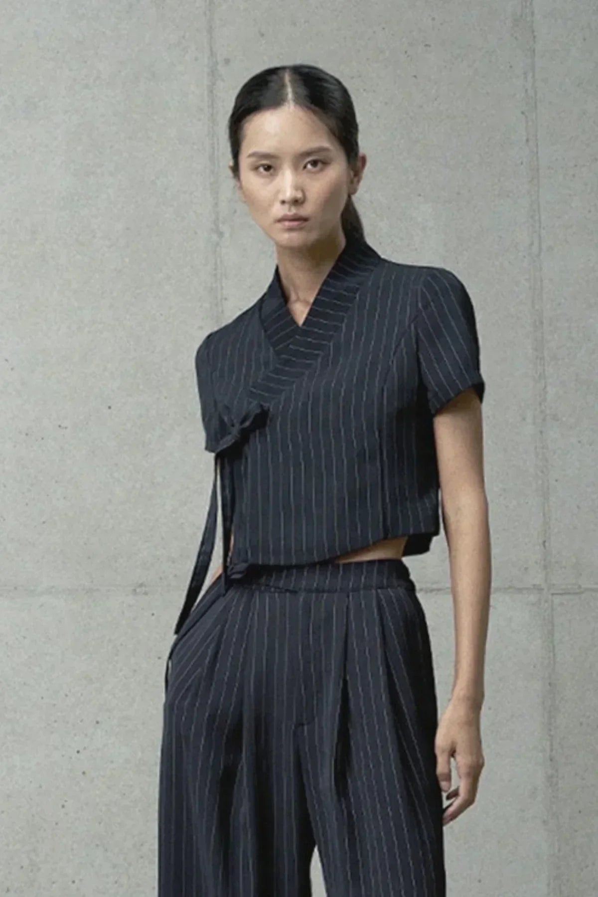 Woman wearing black cropped pinstripe jeogori top with matching high-waisted pants against concrete wall