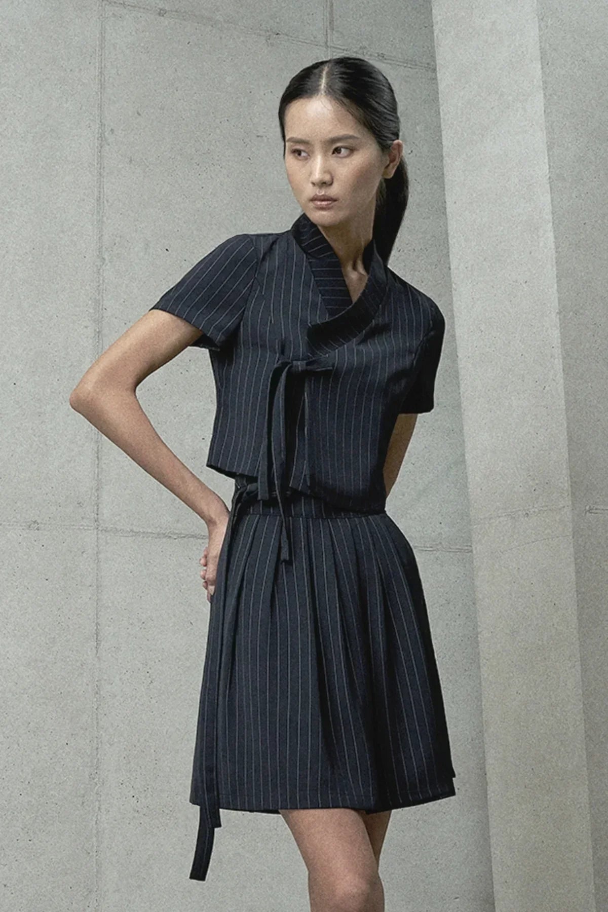 Asian woman wearing a black pinstripe wrap dress with short sleeves against a gray concrete wall