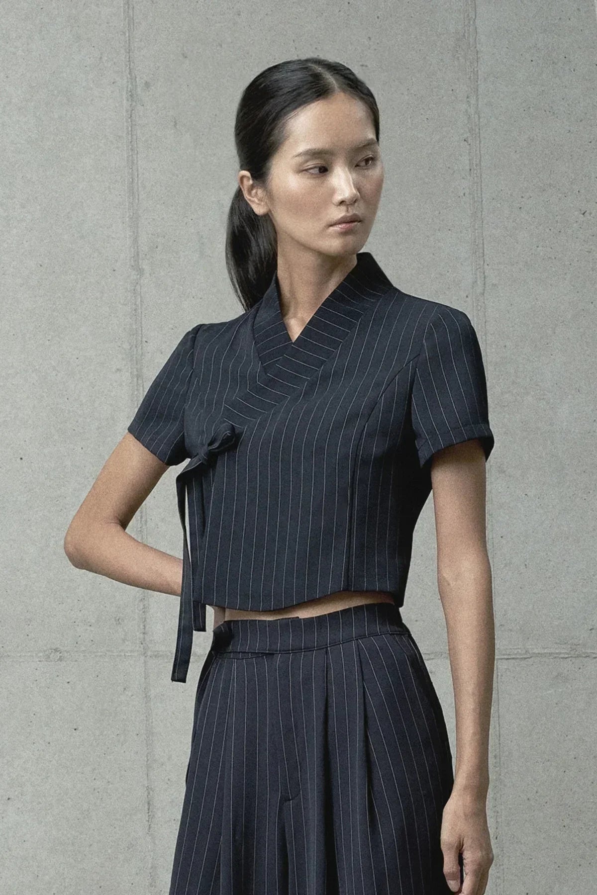 Woman wearing modern black pinstripe short-sleeve jeogori top and matching pants against concrete wall